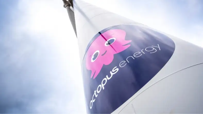 Upward angle view of a wind turbine displaying the Octopus Energy logo and pink octopus mascot on the nacelle