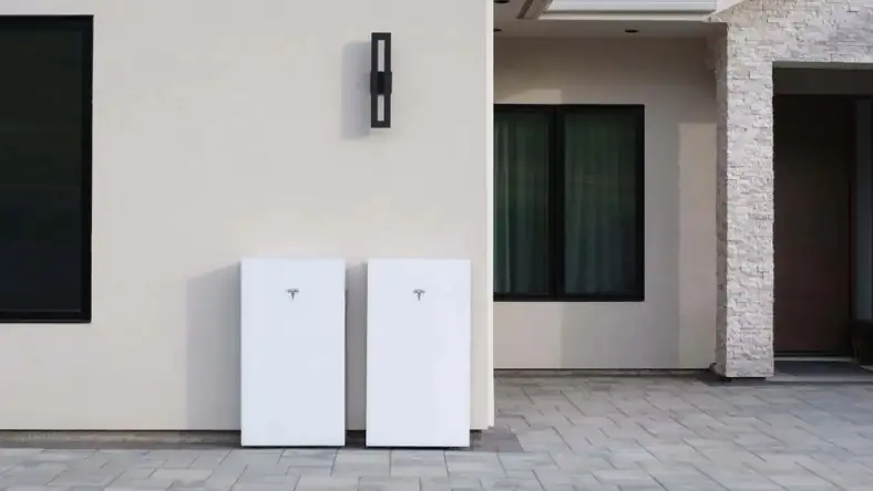 A pair of white Tesla Powerwall home battery units propped outside a modern house with a stone entrance